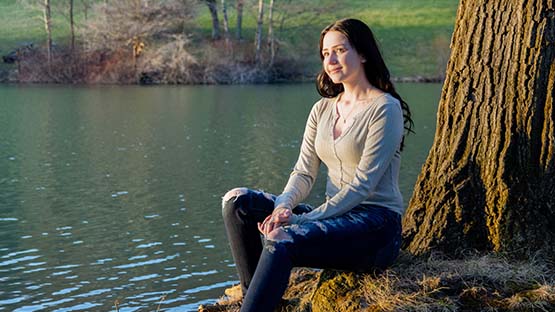 An AHN patient Rachel Wilson sitting on the edge of a body of water next to a tree.