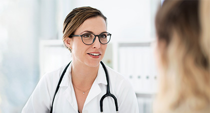 female medical professional wearing glasses with stethoscope around her shoulders speaking to a patient. Image background:  professional office