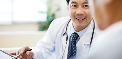 head and shoulders of smiling man wearing a shirt and tie with stethoscope over shoulders leaning forward engagingly with other person who is mostly off image and in soft focus.