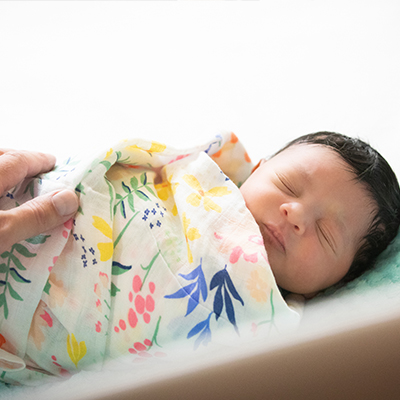 A newborn wrapped in a blanket sleeping in a bassinet with a parent's hand on the baby.