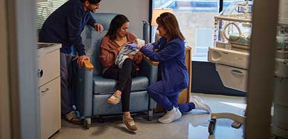 A nurse kneeling next to a smiling mother sitting in a chair holding a newborn with smiling father standing next to them in a hospital room 