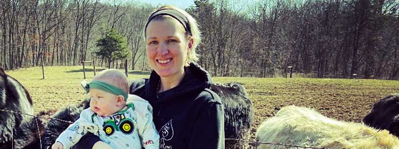 A woman holding a baby in front of a field with trees and some farm animals in the background.