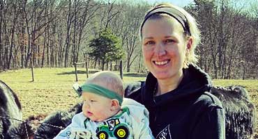 A woman holding a baby in front of a field with trees and some farm animals in the background.