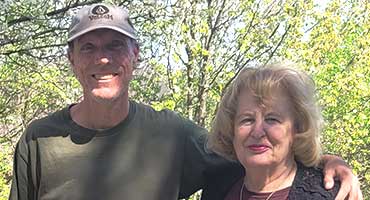 A man in a hat and t-shirt stands with his arm around a woman, both smiling in front of green trees.