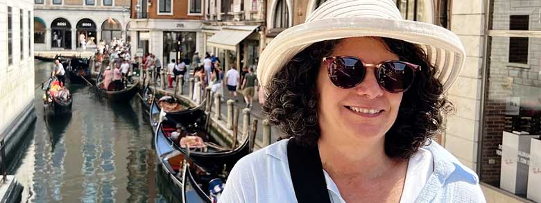 A woman in a striped hat and sunglasses smiles with gondolas and people on a canal in Venice, Italy, behind her.