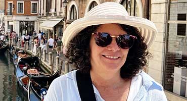 A woman in a striped hat and sunglasses smiles with gondolas and people on a canal in Venice, Italy, behind her.