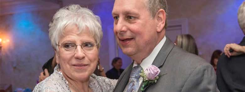 A smiling woman with gray hair and glasses is embracing a man in a suit with a purple boutonnière.