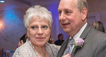 A smiling woman with gray hair and glasses is embracing a man in a suit with a purple boutonnière.