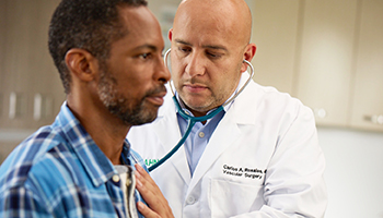 A doctor in an exam room listening to a patient's heart with a stethoscope