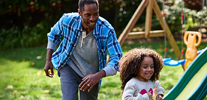 Smiling father chasing daughter around a swingset in backyard