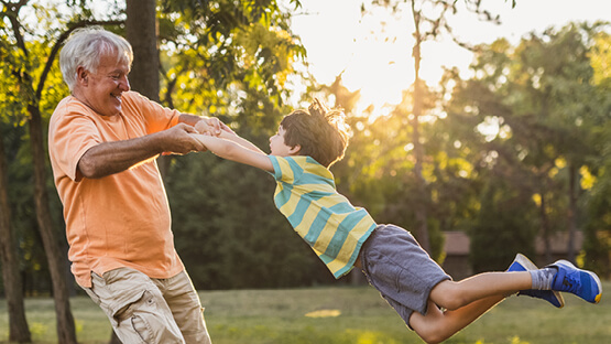 A smiling grandfather spinning his happy grandson outdoors as the sun sets behind the trees