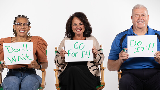Three smiling hip and knee replacement patients sitting in chairs holding hand written signs that say Don't Wait, Go For It, Do it!
