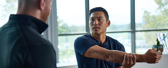 A sports performance patient stretches his right arm across his chest as his physical therapist watches him
