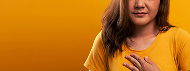 A woman in a yellow dress holding her hand over her chest and have closed her eyes because she is experiencing heart burn.