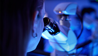 A person looking into a microscope, illuminated by blue light.