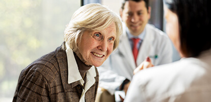 An elderly woman with white hair smiles and gestures while talking to two people.