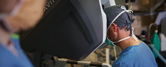 A surgeon wearing a mask and cap looks into a robotic surgical console.