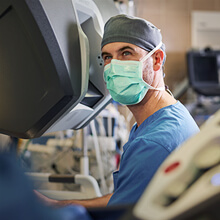 A surgeon in blue scrubs and mask operates a robotic surgical system.