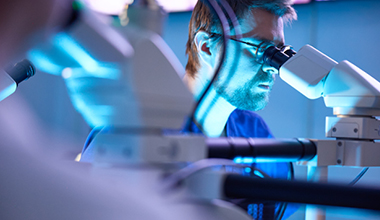 A person with brown hair wears glasses and looks through a microscope.