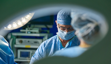 Surgeon wearing glasses, mask, and surgical cap in operating room under surgical lights for cancer care.