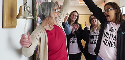 A woman in a pink shirt ringing a bell and celebrating to a cheering crowd of women wearing matching shirts that say She's Outta Here 