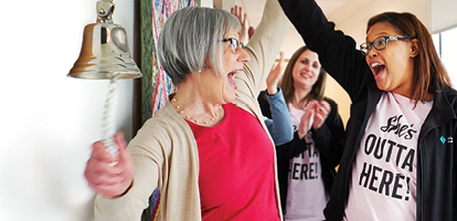 A woman in a pink shirt ringing a bell and celebrating to a cheering crowd of women wearing matching shirts that say She's Outta Here 