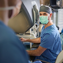 A white medical professional in blue scrubs and a mask operates equipment.