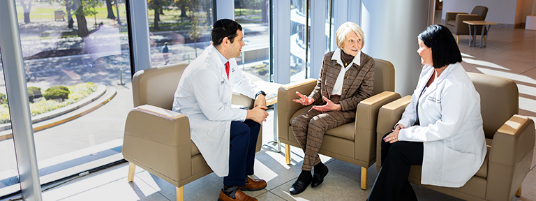 Two medical professionals and an older woman are talking in a sunny waiting room.