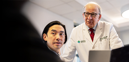 A white medical professional in a lab coat and tie speaks to an Asian man in a suit.