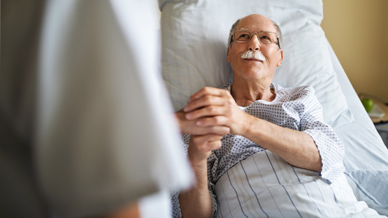 An elderly man with glasses and a white mustache laying in a hospital bed and looking at a doctor that he is shaking hands with.