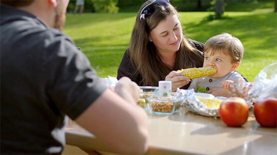 A mom and dad having a picnic with their young son and the mom is feeding the son corn.
