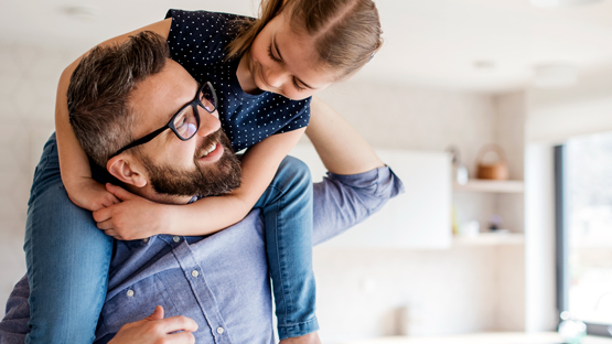 A father wearing a dress shirt and glasses with his daughter on his shoulder who is hugging him around the neck..