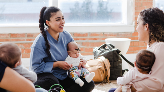 Three mothers sitting on the floor in a bright room talking while holding their infants in their laps