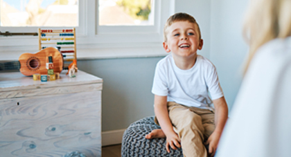 A young boy smiling seen over a doctor's shoulder while sitting a room with toys visible on a chest nearby