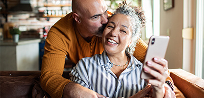 A man kisses a smiling woman's cheek while she holds a smartphone.