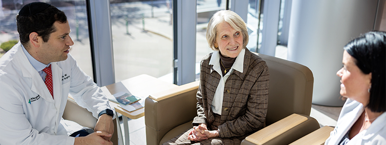 A medical professional in a white coat chats with a smiling woman while another medical professional listens.