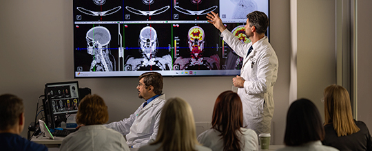 A group of medical specialists are in a room while a doctor is pointing at a large screen of x-ray scans.