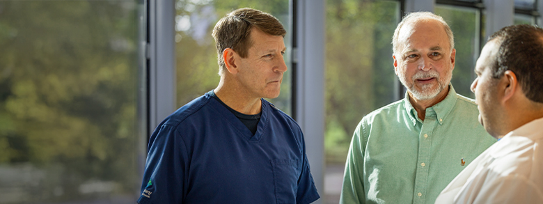 A medical professional in blue scrubs talks with a dark haired man and a person with gray hair and beard.