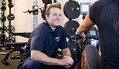 A medical professional in a dark polo smiles at a patient in a gym.