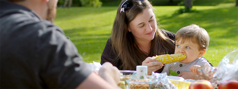 women sitting outside at table with family feeding corn on the cob to her infant